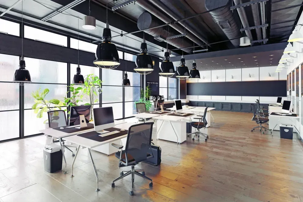 Modern office space with desks, computers, chairs, and overhead lighting. Windows and plants.