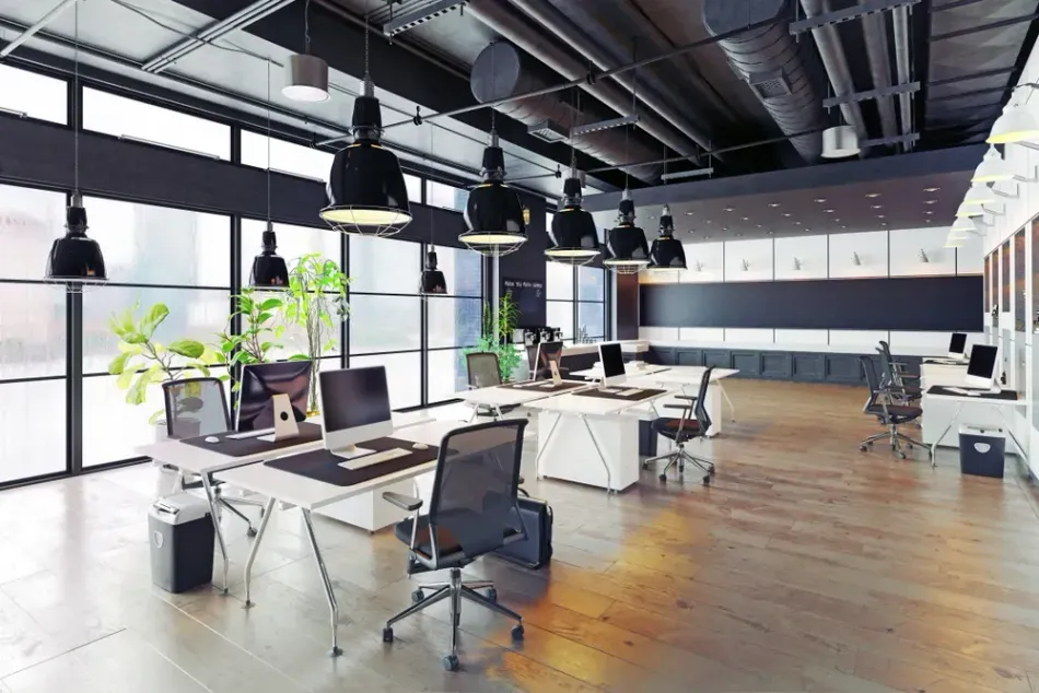 Modern office space with desks, computers, chairs, and overhead lighting. Windows and plants.