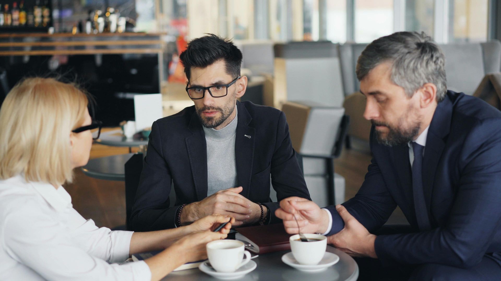 Three people in business attire discuss over coffee at a cafe table.