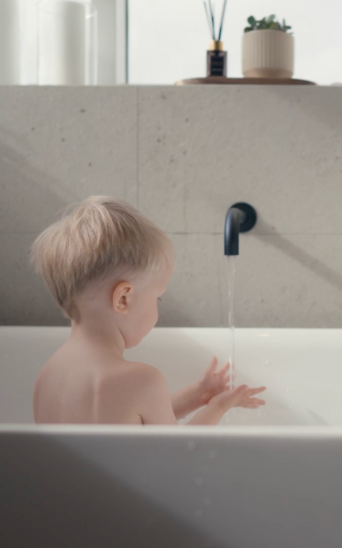 Young child in a bathtub washing hands under a black faucet in a bright bathroom