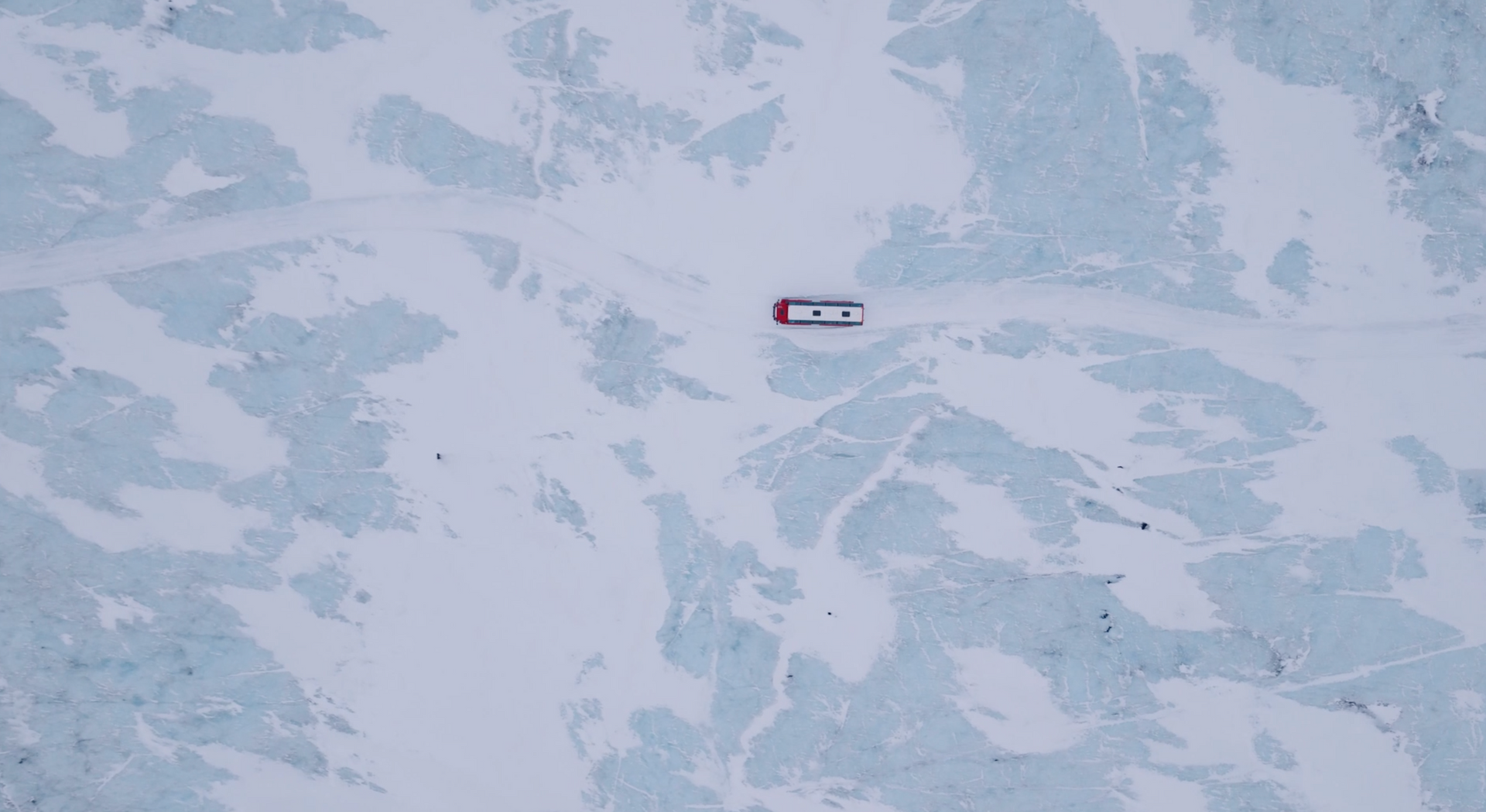 Small red-and-white vehicle crossing a snowy, icy landscape seen from above.