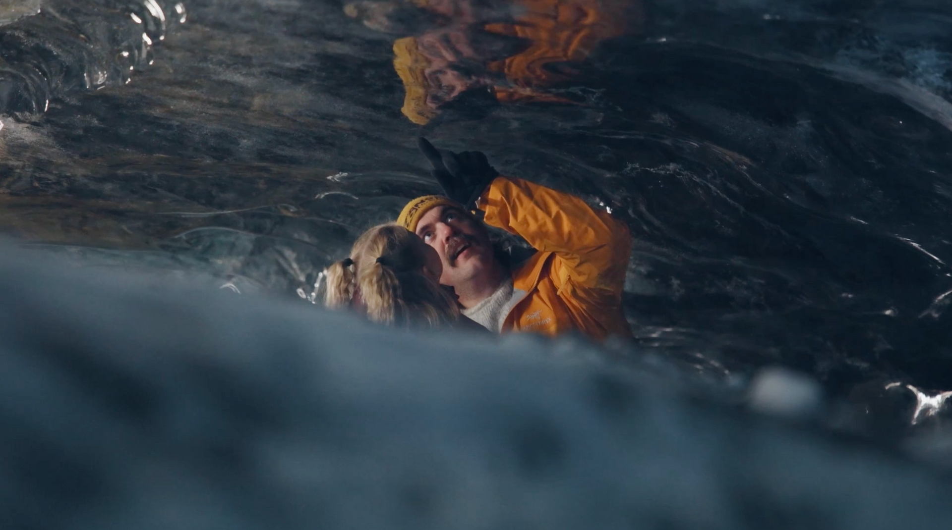 Two people embrace in rough water beneath a dark cave ceiling