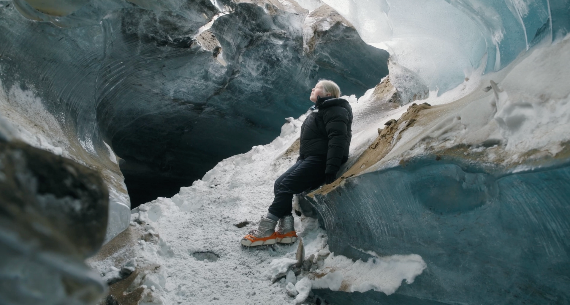 Person sitting in a blue ice cave beside a snow-covered opening, wearing dark winter gear and orange boots
