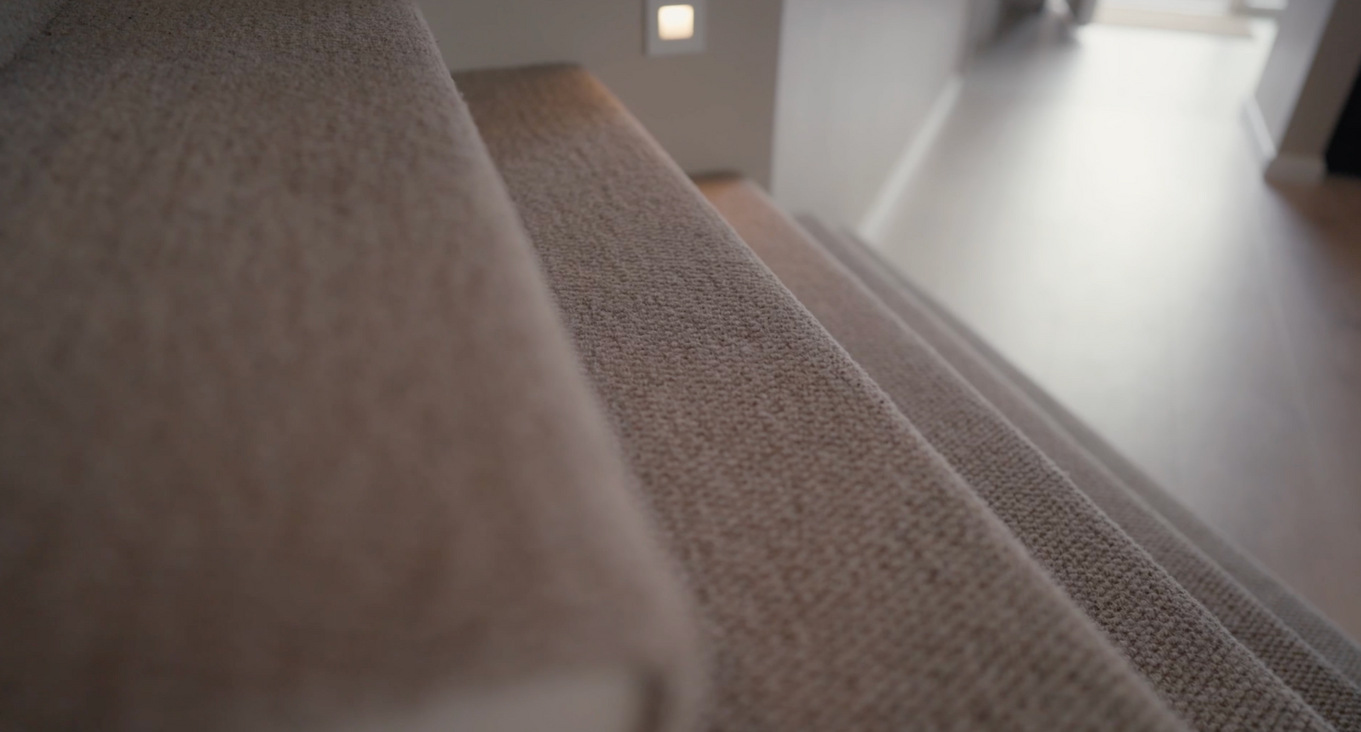 Close-up of a carpeted stair edge beside a blurred hardwood floor in a bright room
