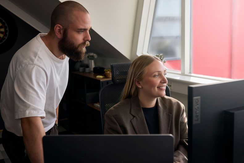 Two coworkers smiling at computer monitors in a bright office