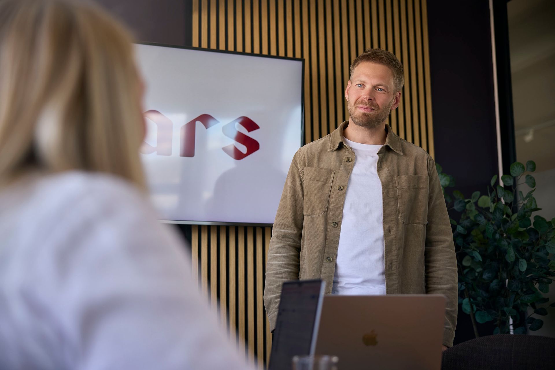 Person standing in a meeting room beside a laptop, facing an audience with a presentation screen behind them