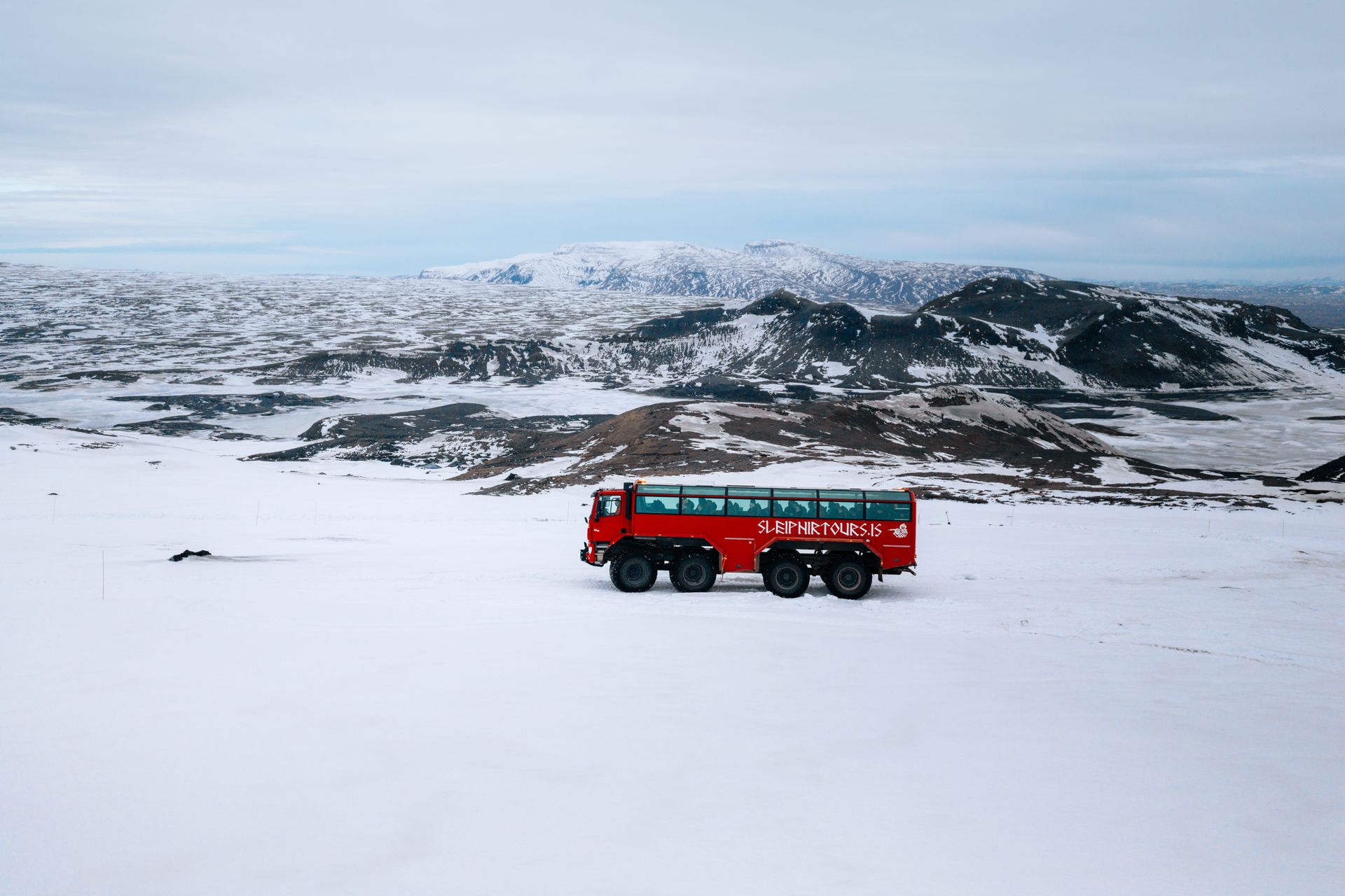 Red bus driving across a snowy plain with mountains in the background