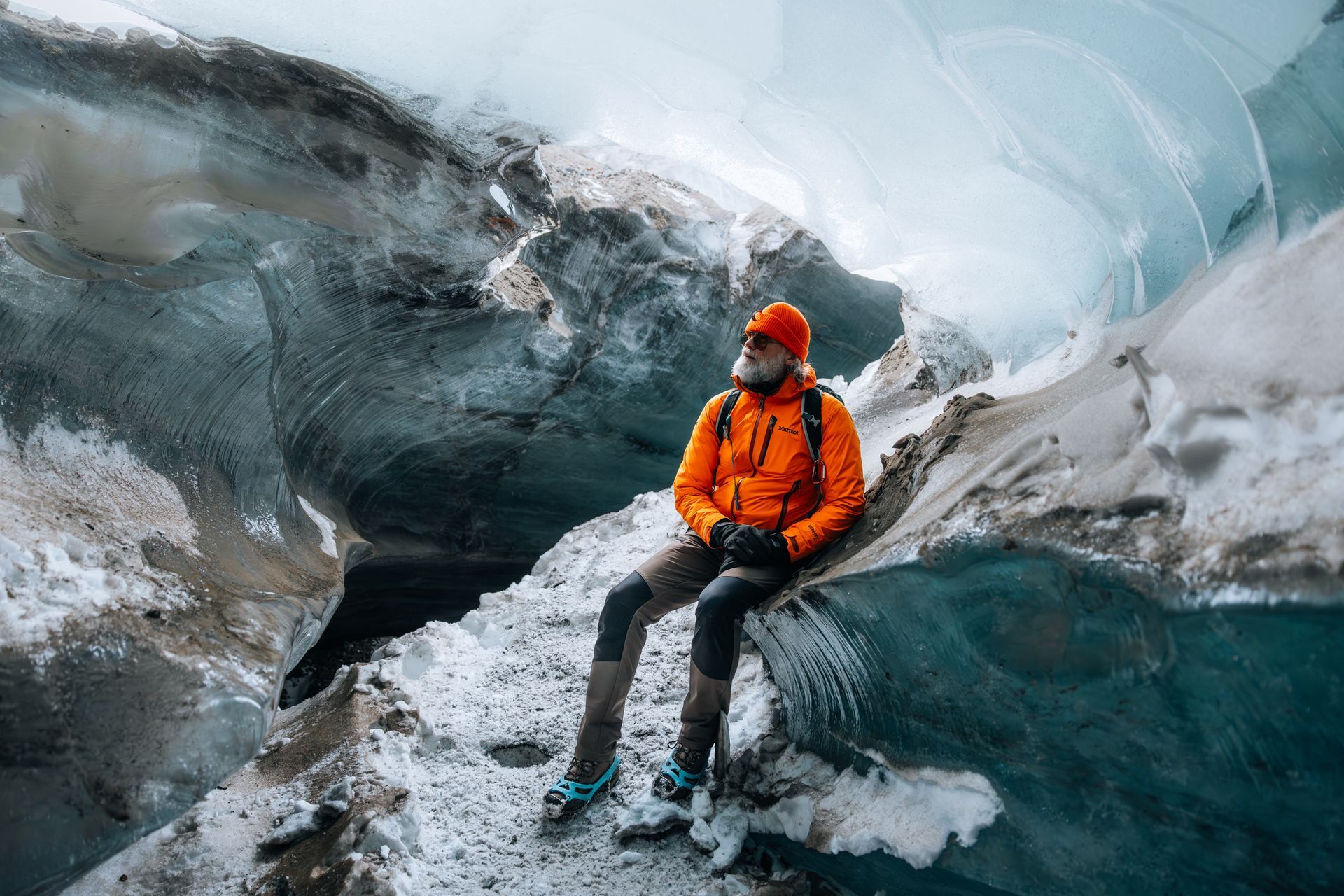 Person in orange jacket sitting on an icy cave ledge inside a blue glacier.