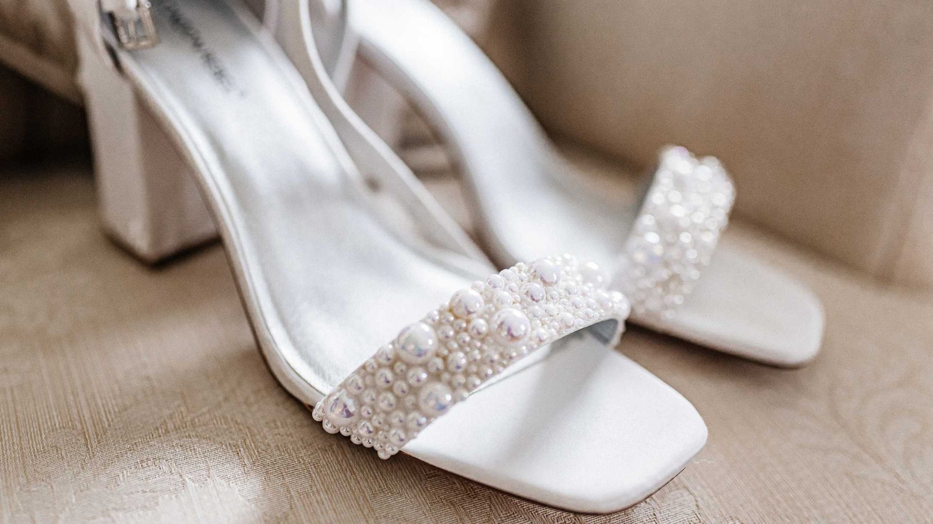 A pair of white wedding shoes with pearls on them are sitting on a couch.