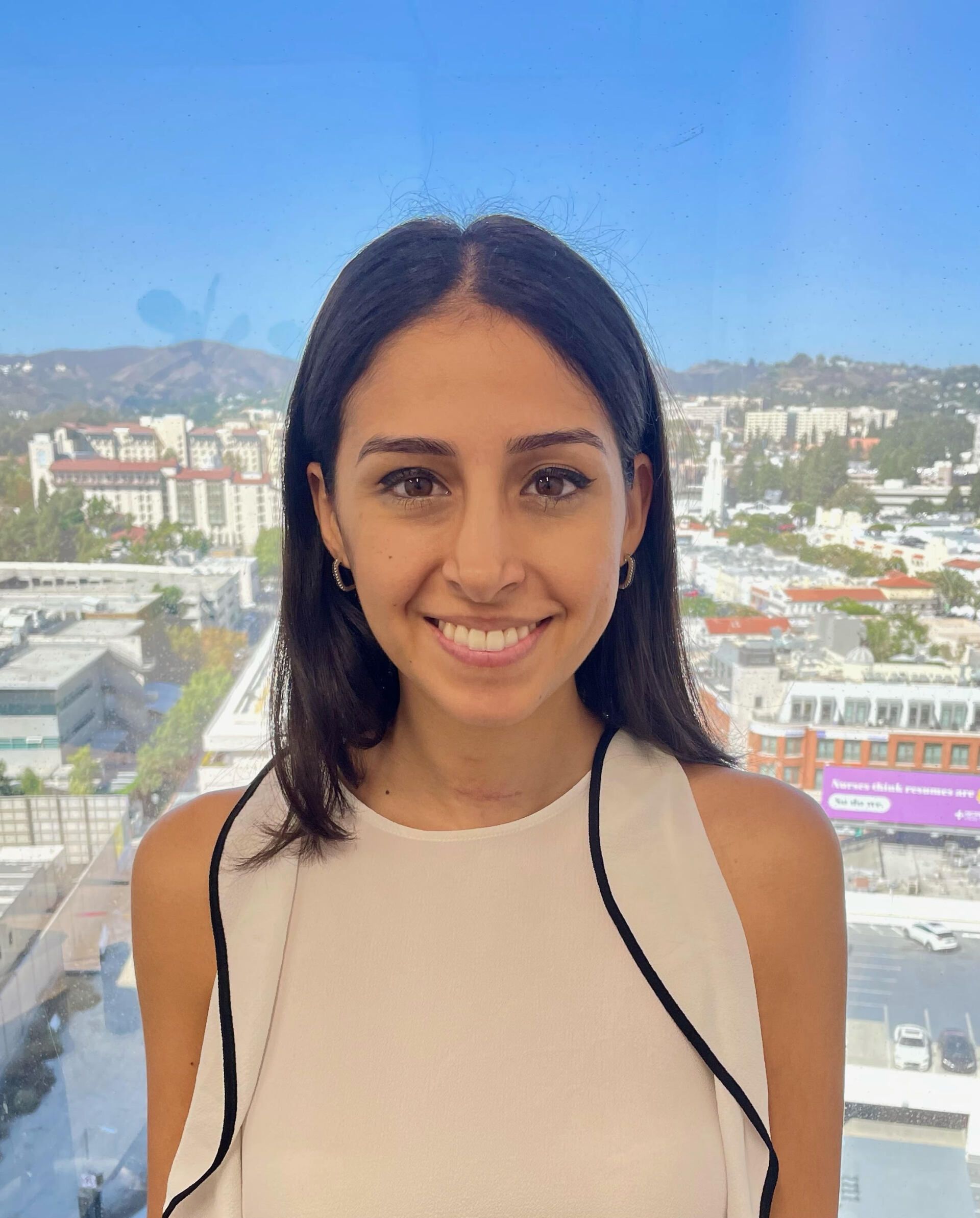 A woman in a white tank top is smiling in front of a city skyline.