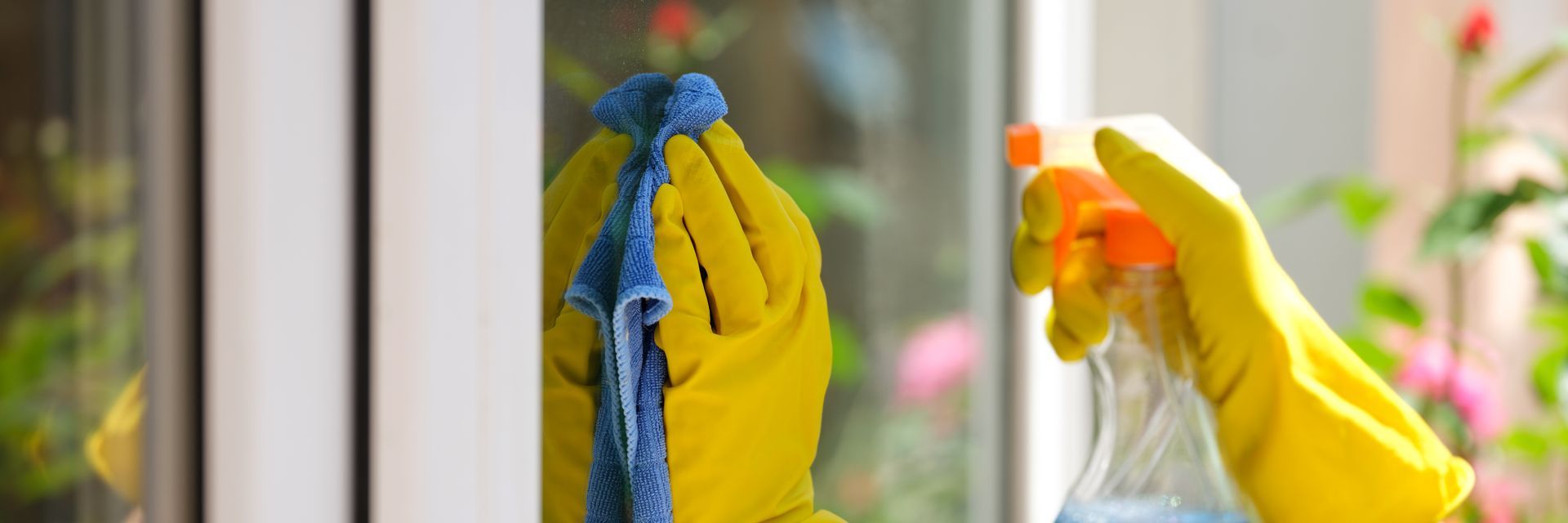 A person wearing yellow gloves is cleaning a window with a cloth and spray bottle. There are spring flowers in the background.