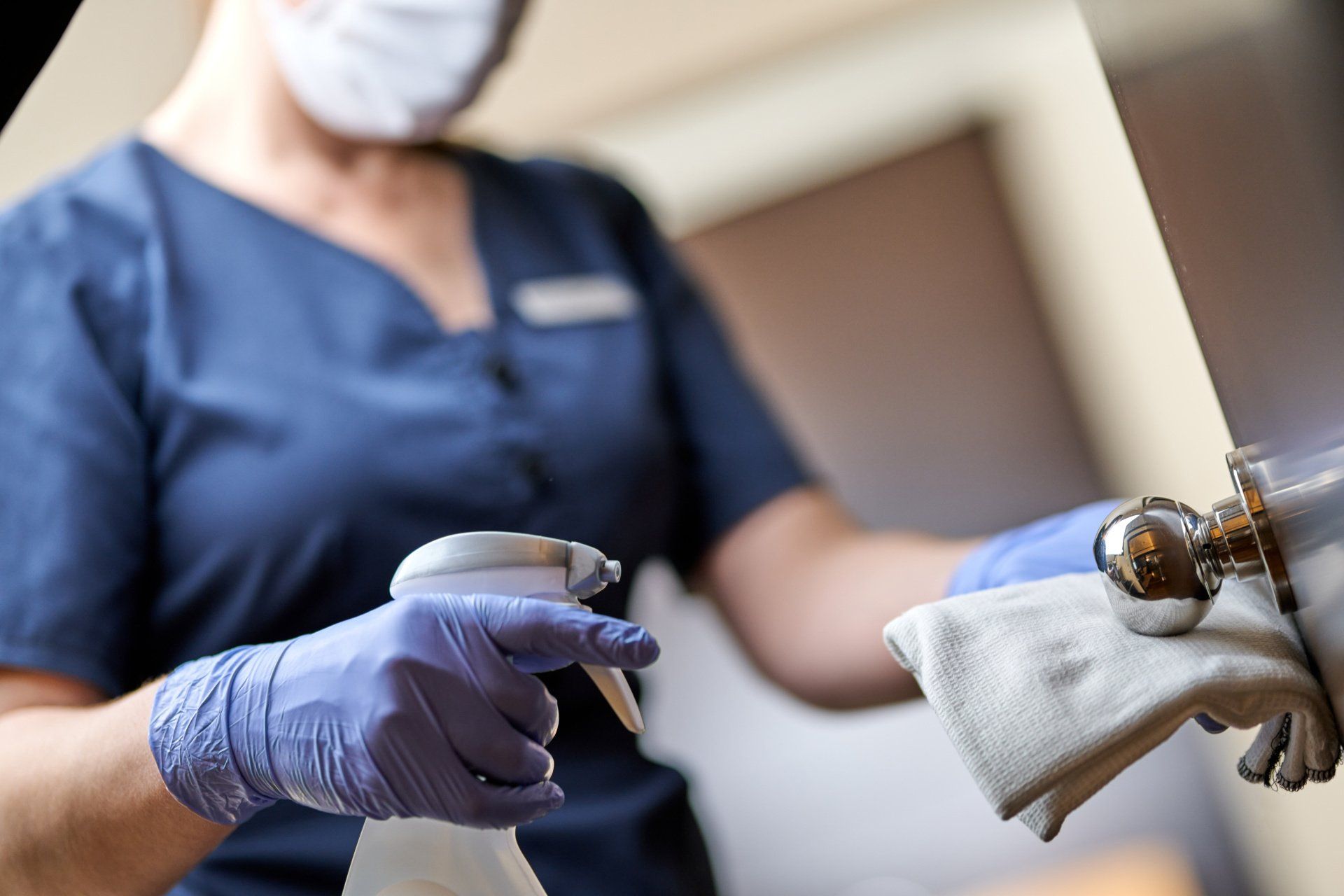 A woman wearing a mask and gloves is cleaning a door handle with a cloth.