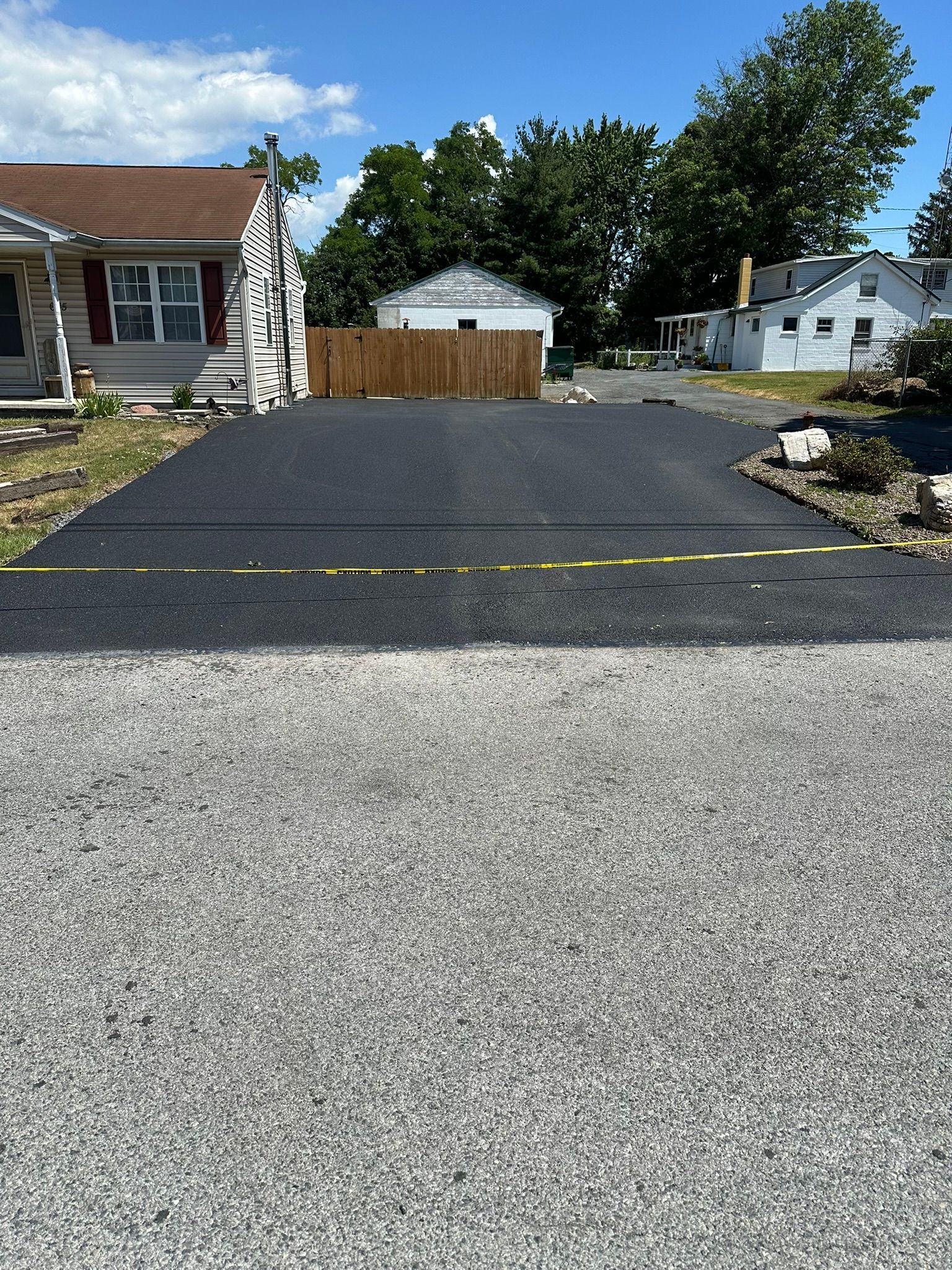 Fresh black asphalt driveway in a residential neighborhood, leading to a wooden garage under blue sky.