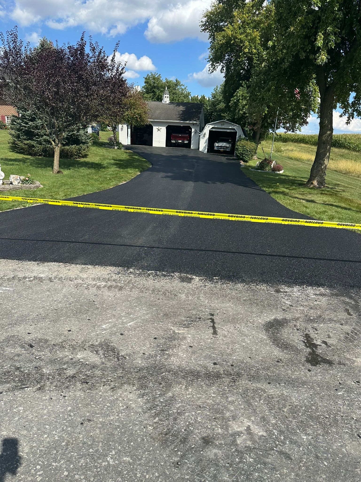 Driveway leading to a garage with a black car, bordered by trees and a yellow caution tape across the entrance
