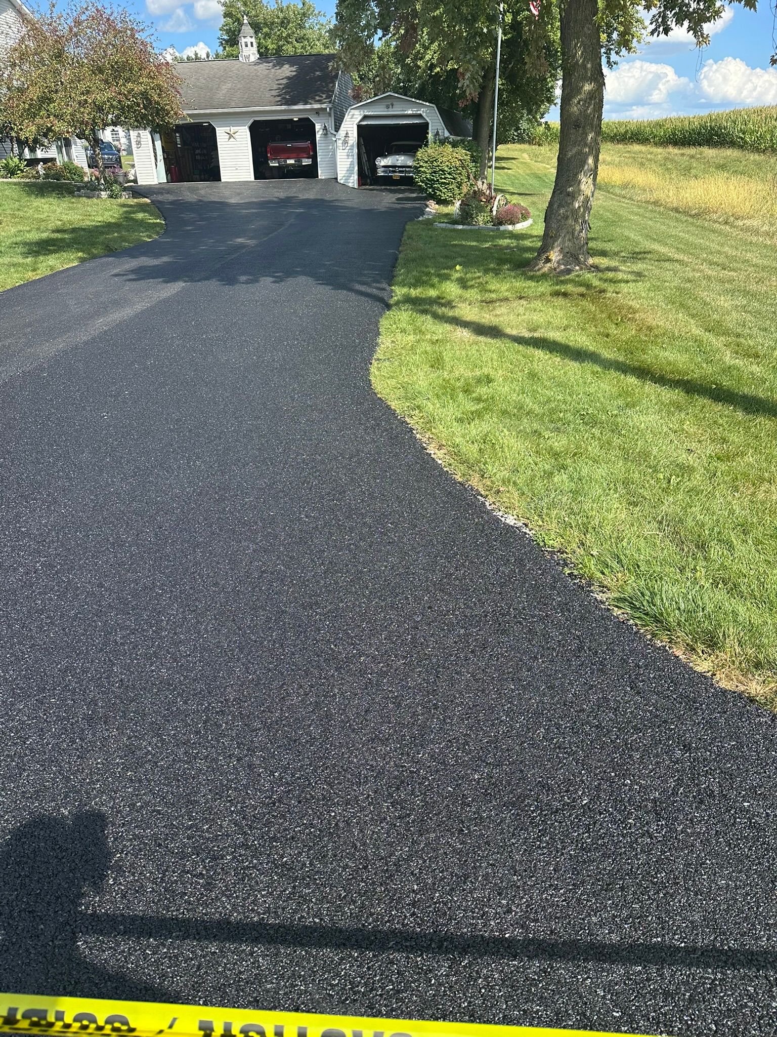 Fresh black asphalt driveway leading to a garage beside a green lawn and trees on a sunny day