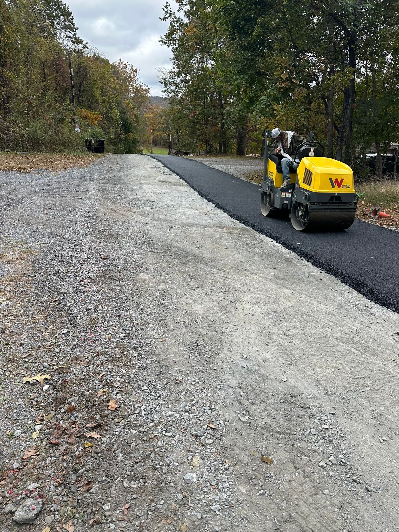 Construction worker compacting fresh asphalt on a gravel road beside trees in autumn