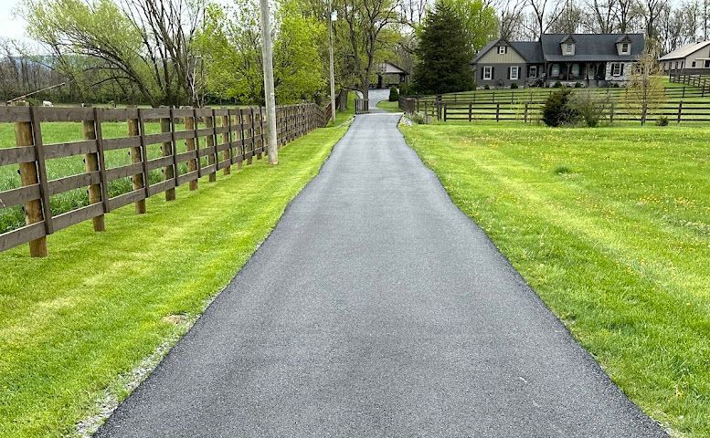 Paved driveway through green lawn to a house, with a wooden fence along the left side.