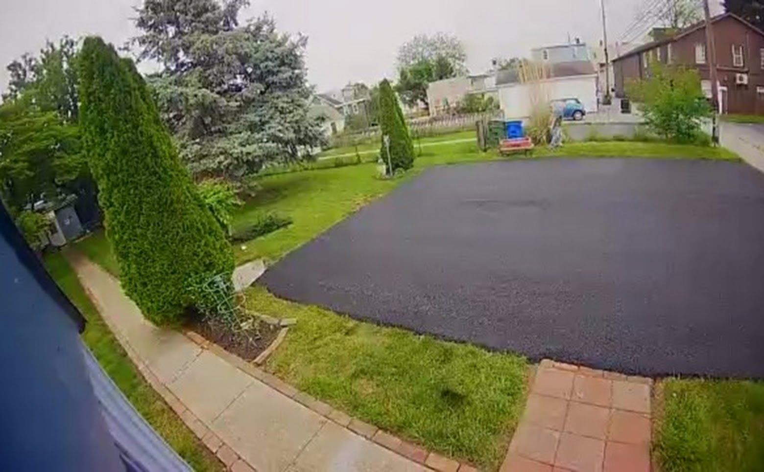 Rainy suburban driveway and front yard with wet asphalt, green lawn, sidewalk, and houses in the background