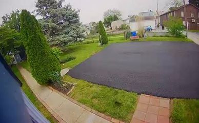 Rainy suburban driveway and front yard with wet black asphalt, green grass, trees, and houses in the background