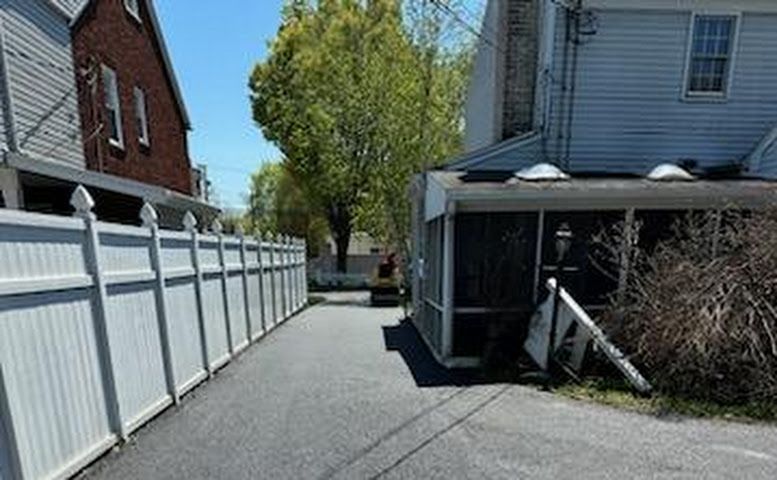 Narrow paved alley between houses with a white fence and a porch, lined by trees in the background