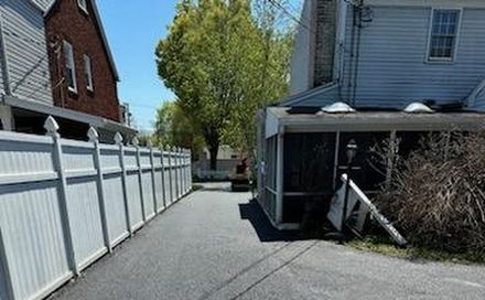 Narrow paved side yard beside a house, with a white fence and trees at the end.