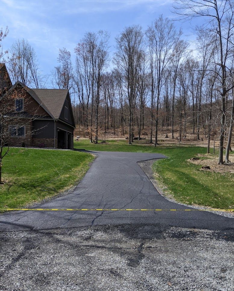 Driveway leading to a brick house beside a wooded yard with leafless trees under a blue sky