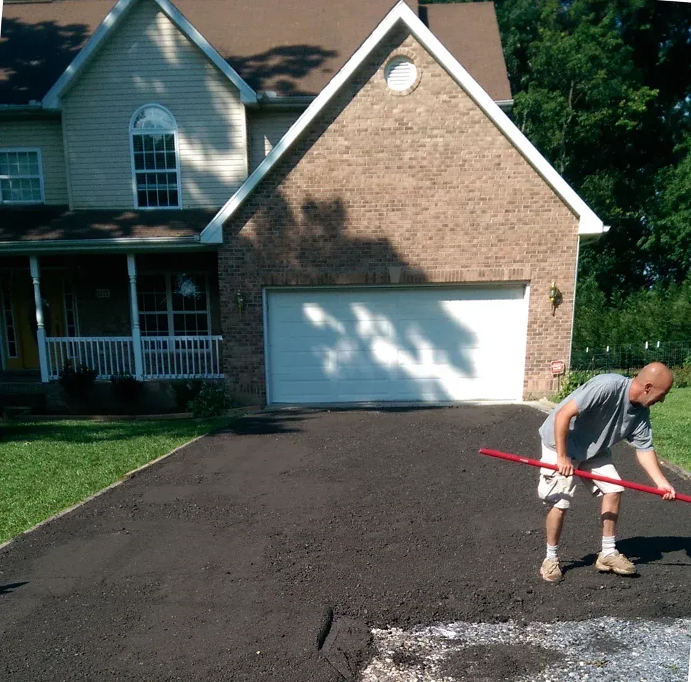 Person raking a driveway beside a suburban house with a garage.
