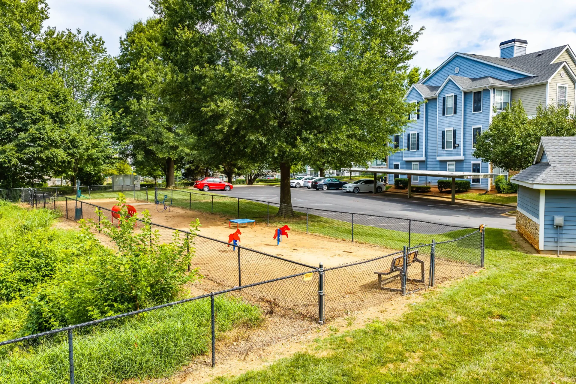 Dog park with play equipment and seating, adjacent to apartment buildings and parking.