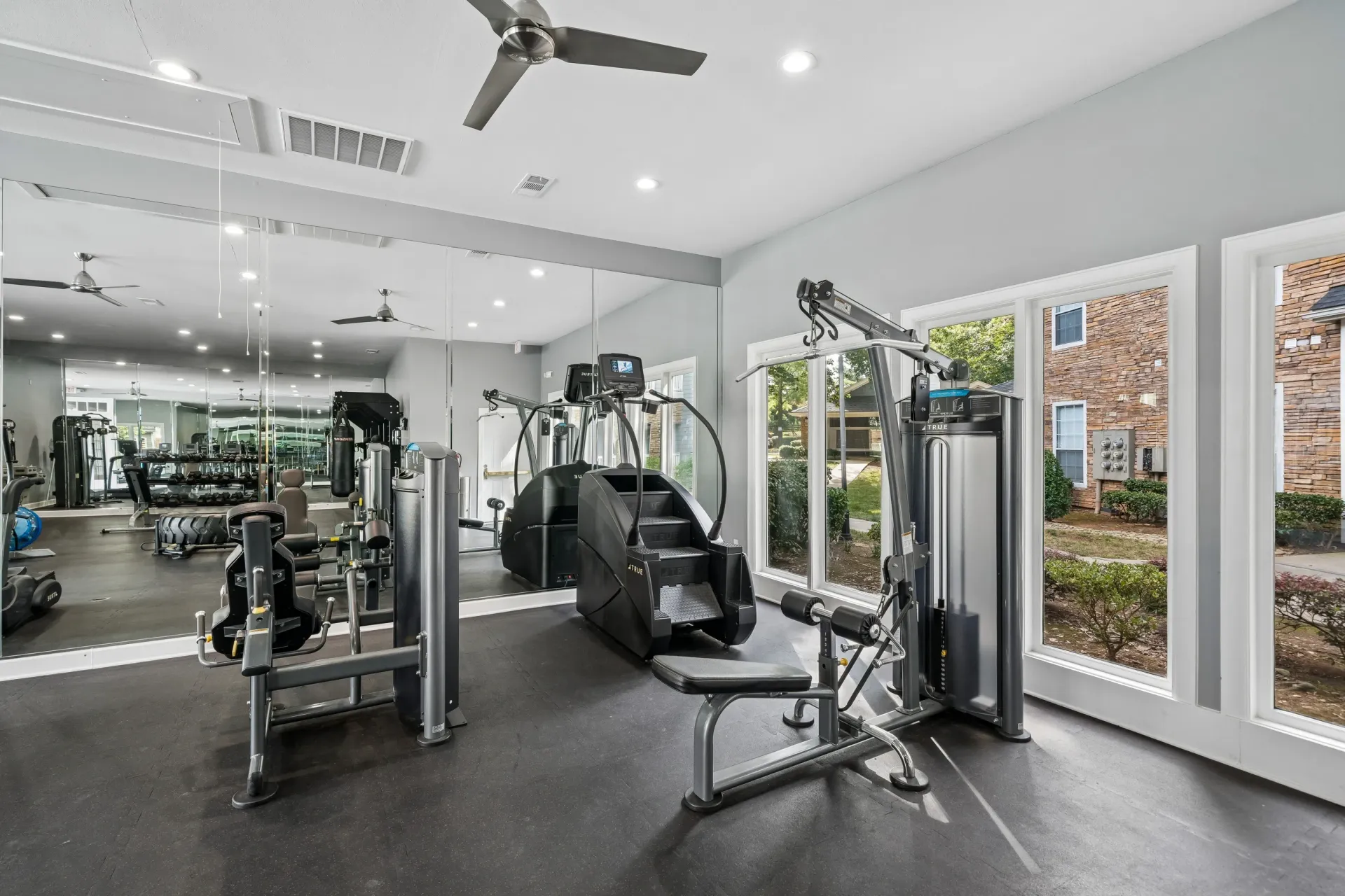 Gym equipment including weight machines and a stair climber, with mirrors on the wall.