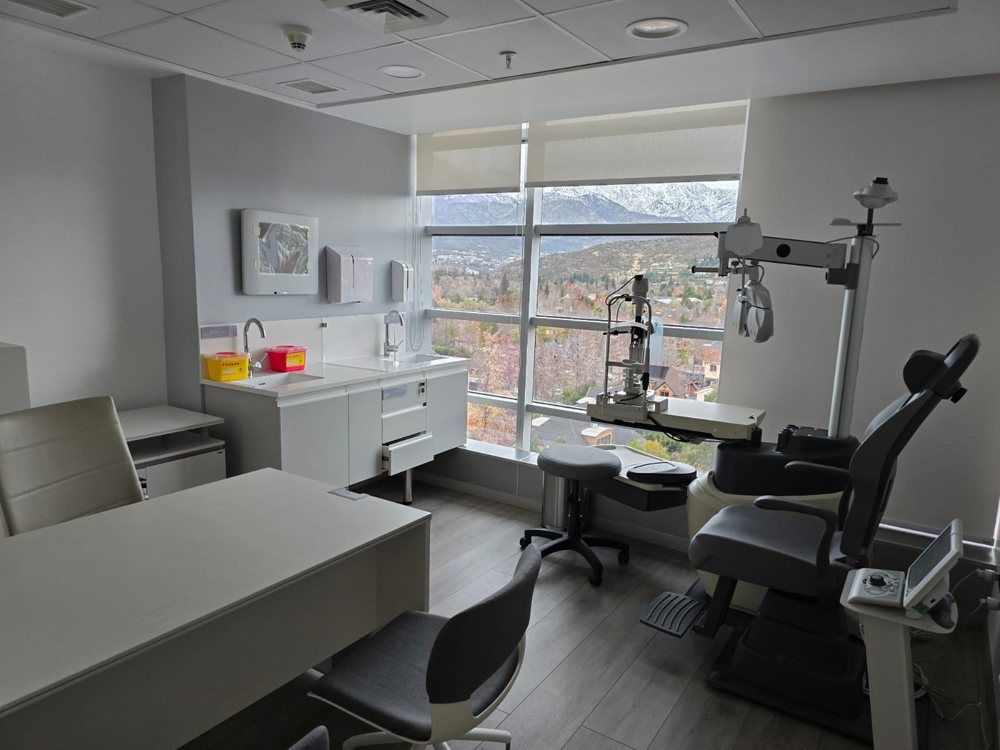Sala de examen dental con sillón, luz de techo, armarios y gran ventanal con vistas a los edificios.