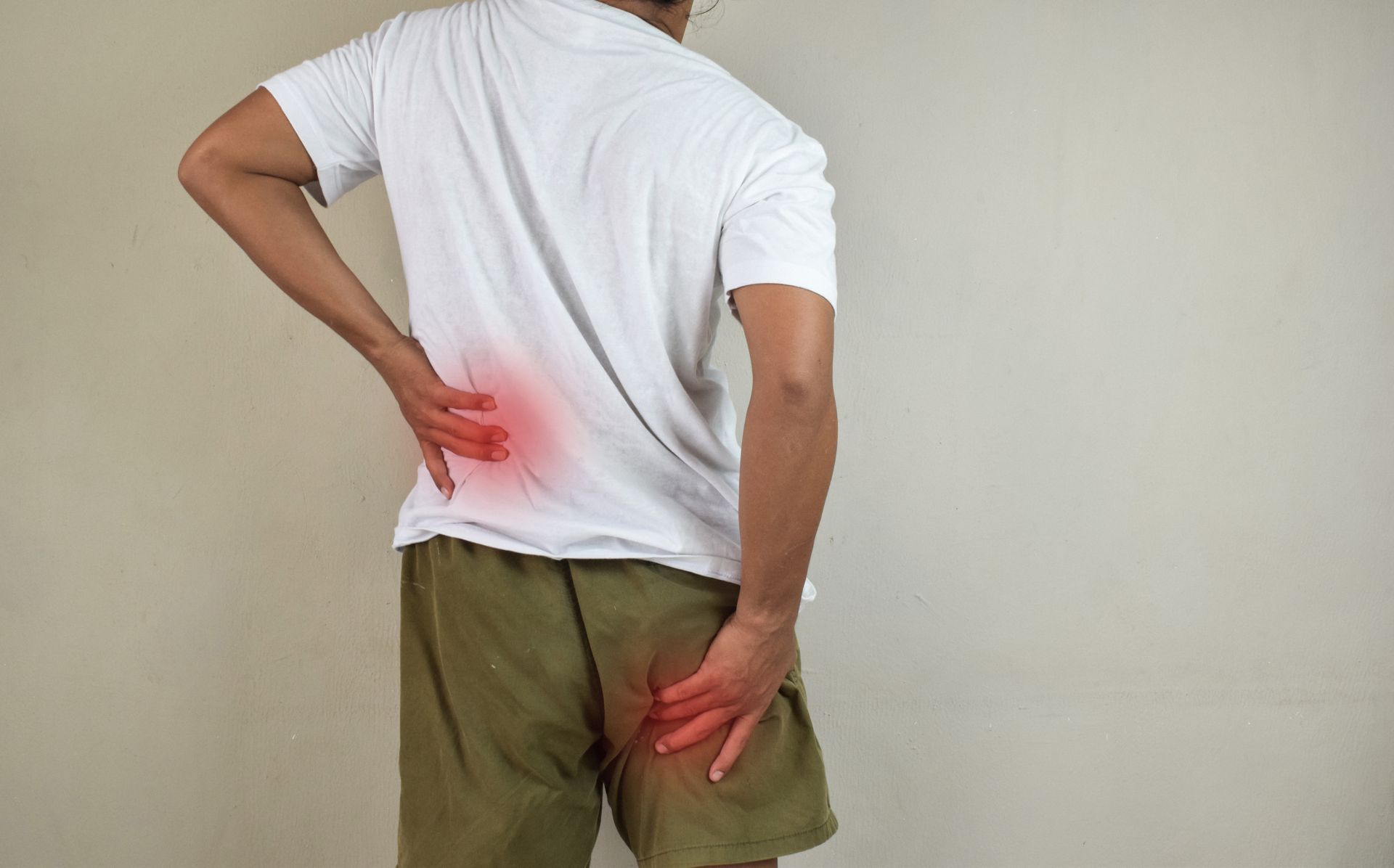 A person in a white shirt and shorts holding their lower back and glute, with red highlights showing pain locations.