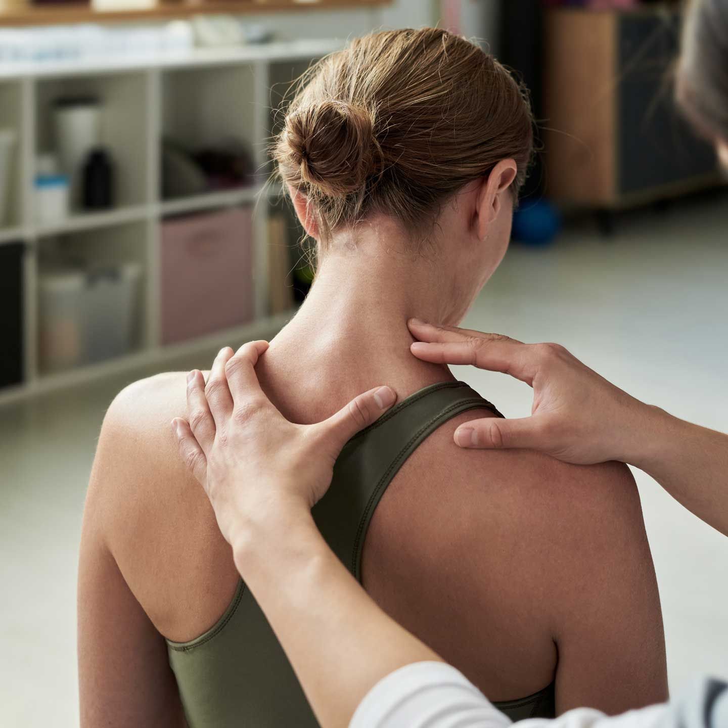 Person receiving shoulder and neck massage indoors. A physical therapist examines the shoulder and neck area of a person wearing an olive green tank top in a clinic.