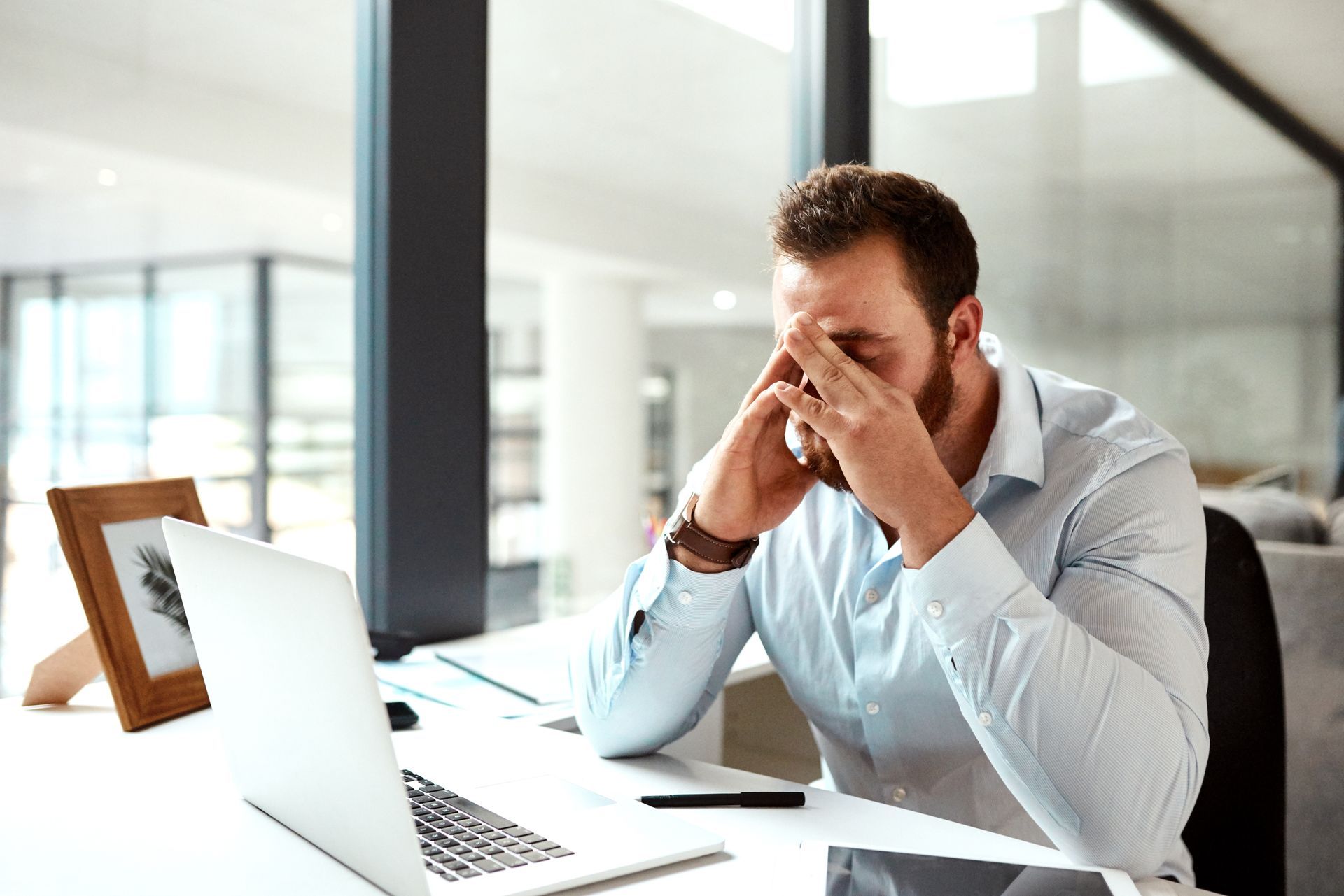 A person sitting at a desk with a laptop, head in hands with a frustrated or stressed expression in an office setting.