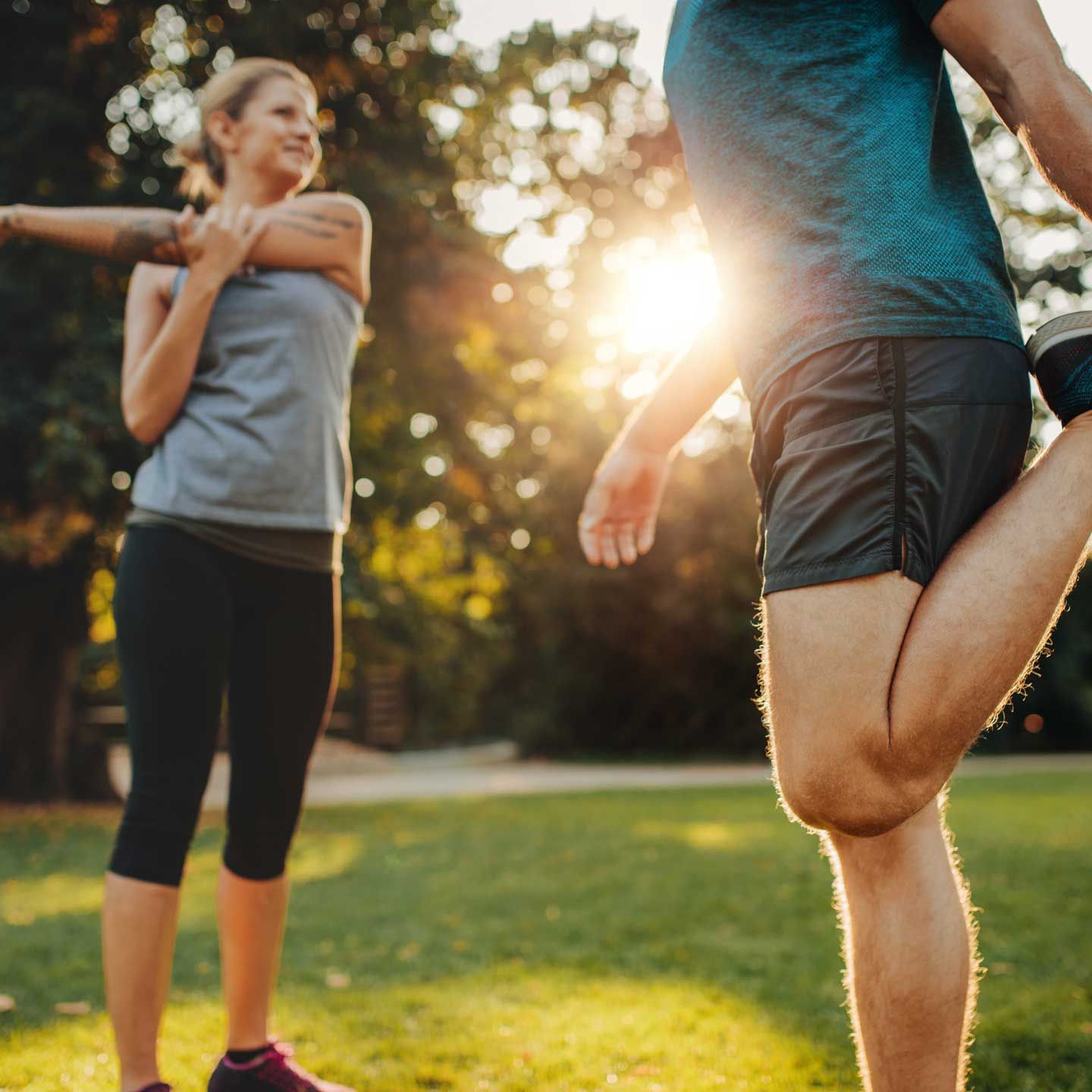 A person stretches their arm across their chest while another person stretches their quadriceps in a sunlit park.