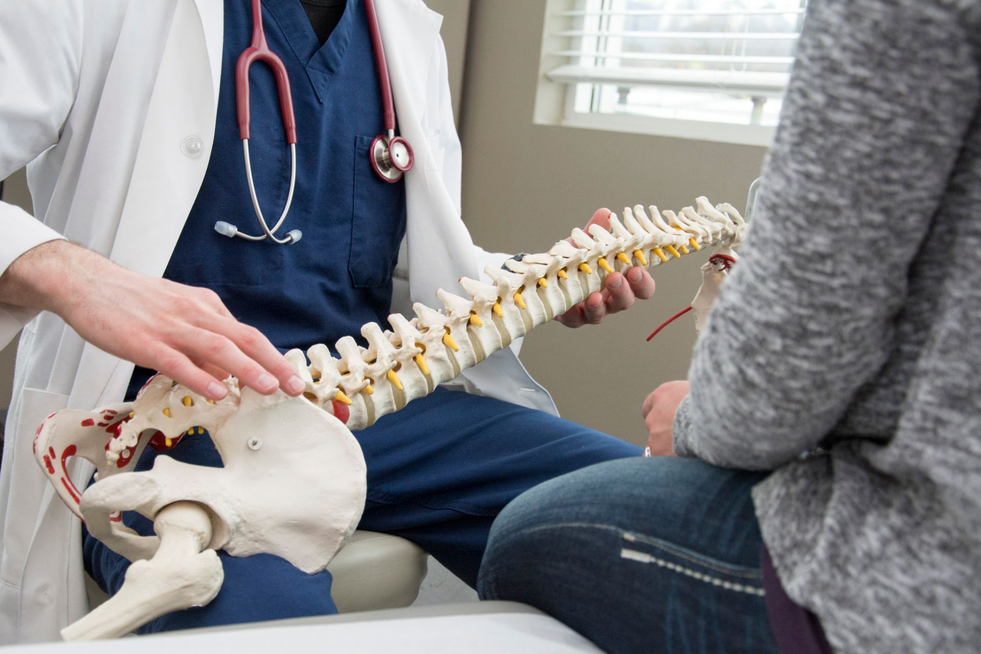 A healthcare provider in a lab coat and stethoscope points to a model of a human spine while consulting with a patient.
