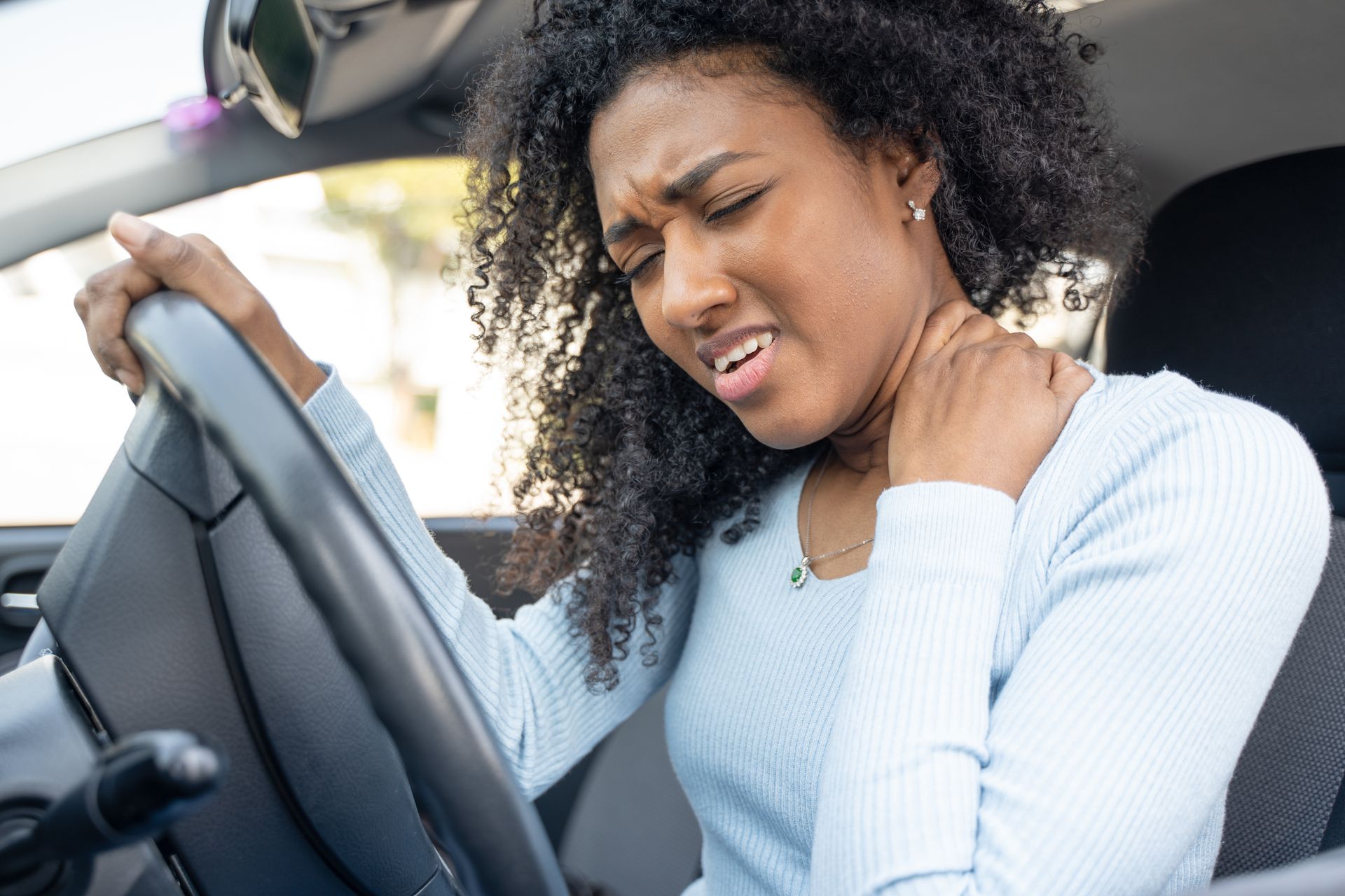 A person in a car, holding their neck in pain with a grimacing expression while sitting at the steering wheel.