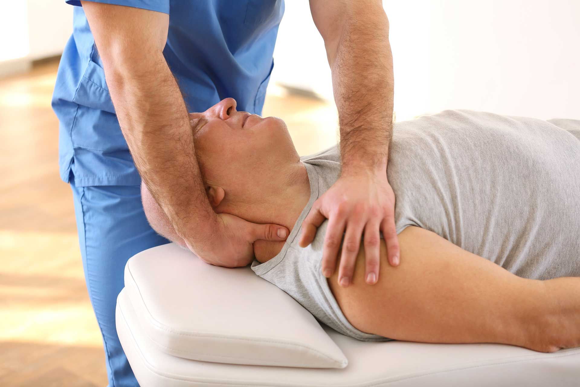 Person receiving neck adjustment from medical professional in clinic. A practitioner in blue scrubs performs a neck adjustment on a patient lying on a treatment table.