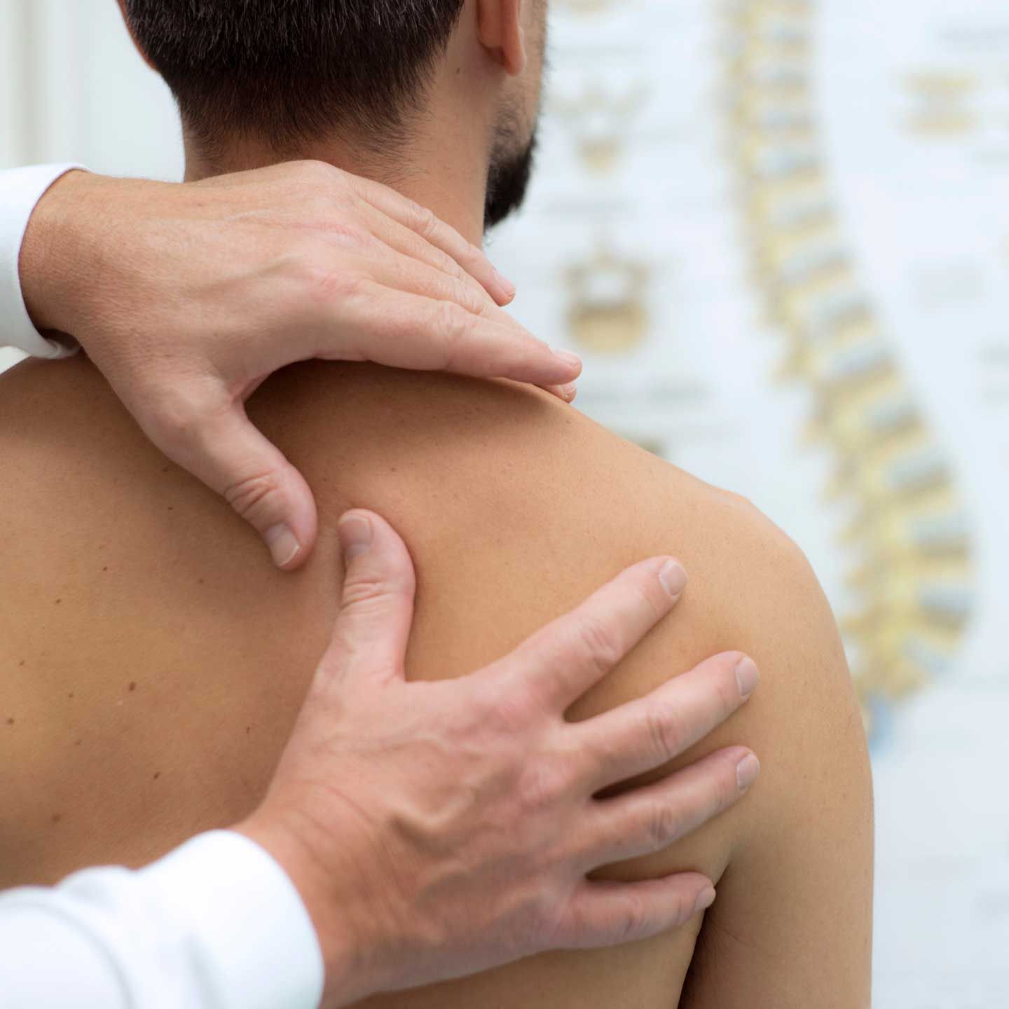 Hands of a healthcare professional examining a person's shoulder, with a spine chart in the background. A healthcare professional performs a physical assessment on a patient's upper back near a spine anatomy chart.