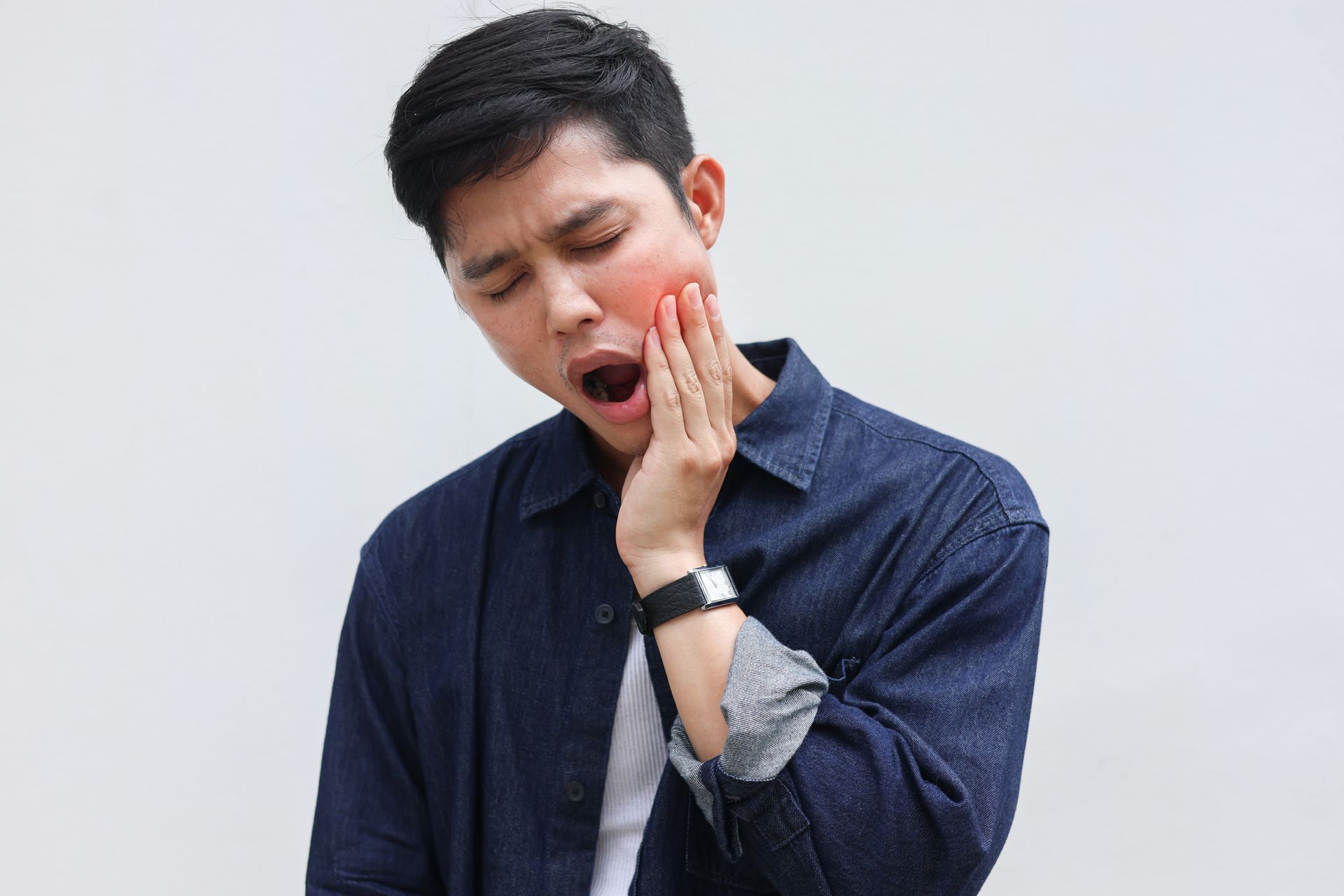 A person in a denim shirt holding their cheek with an expression of pain, indicating a toothache.