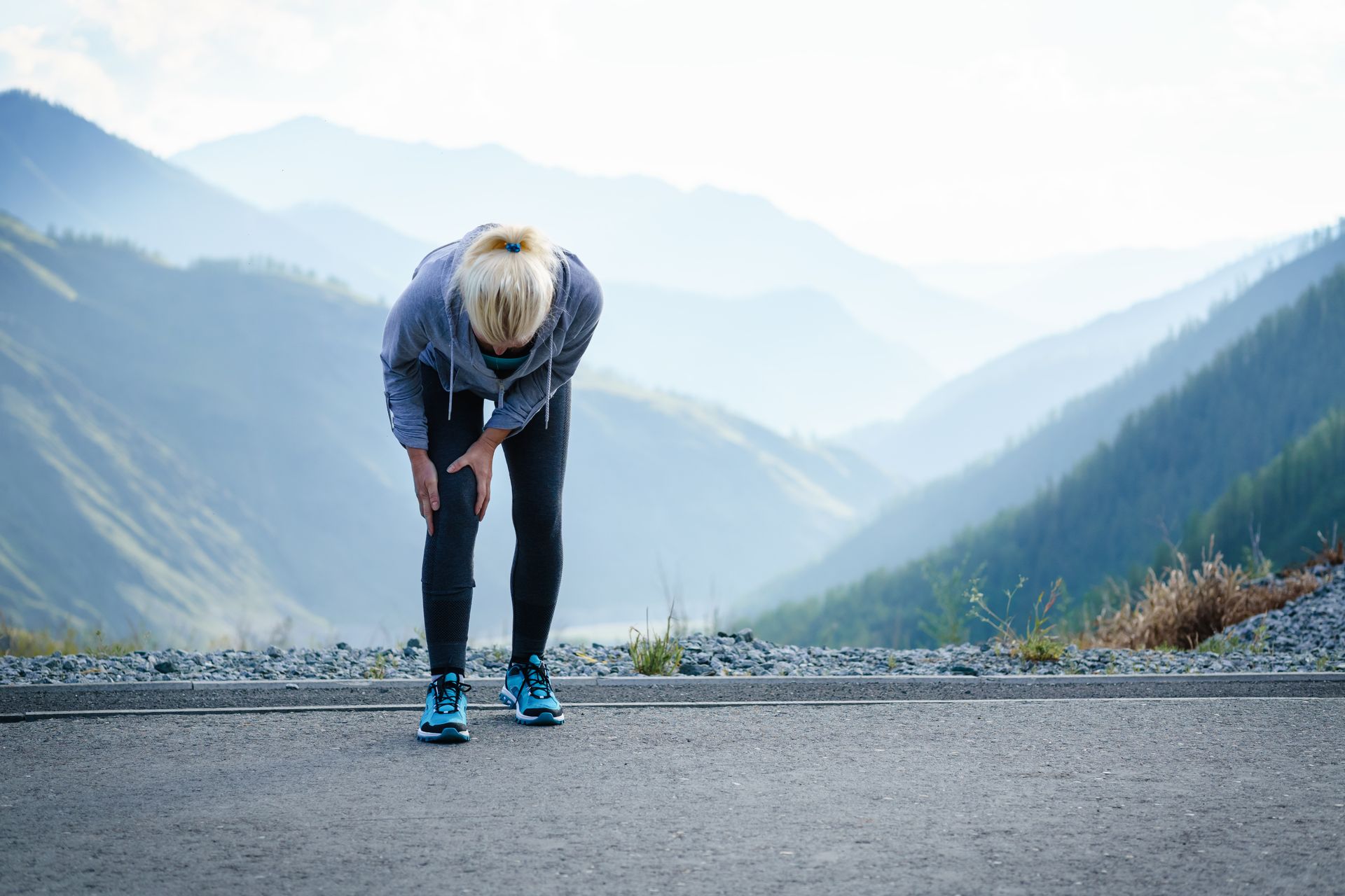 Doctor examining patient's back, palpating the lower back area. A person in athletic gear stands on a mountain road, bent over and holding their knee in apparent pain.