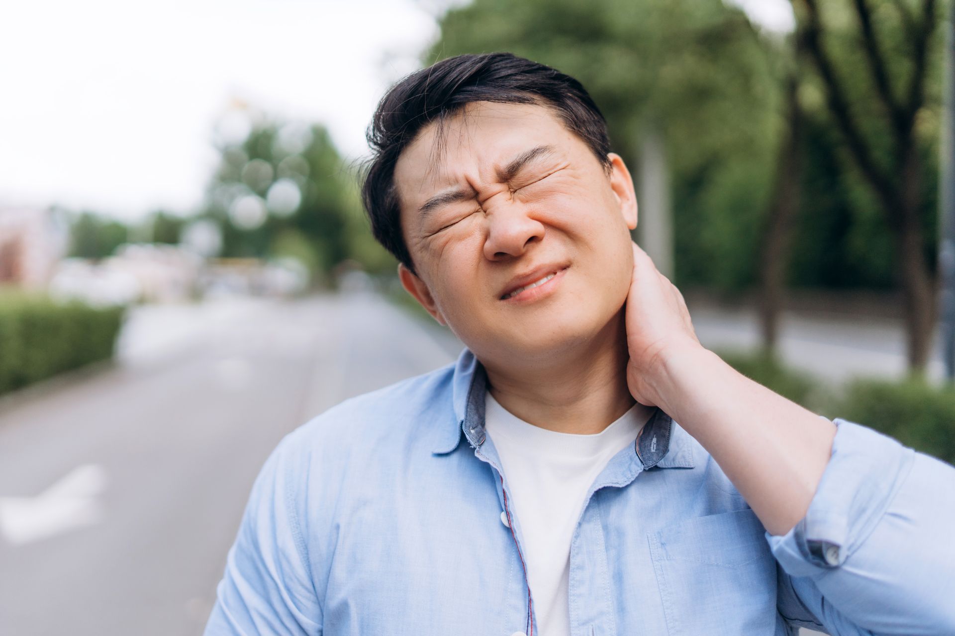 A person in a light blue shirt outdoors, holding the back of their neck with an expression of pain or discomfort.
