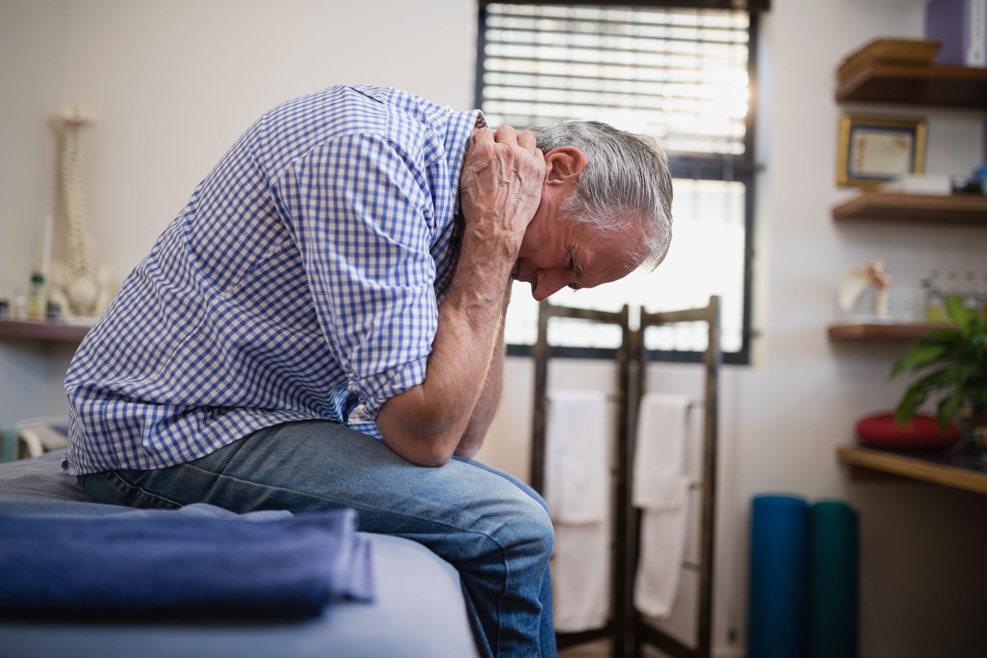 A person sits on a therapy table, hunched over with their hands clasped behind their neck in a clinic setting.