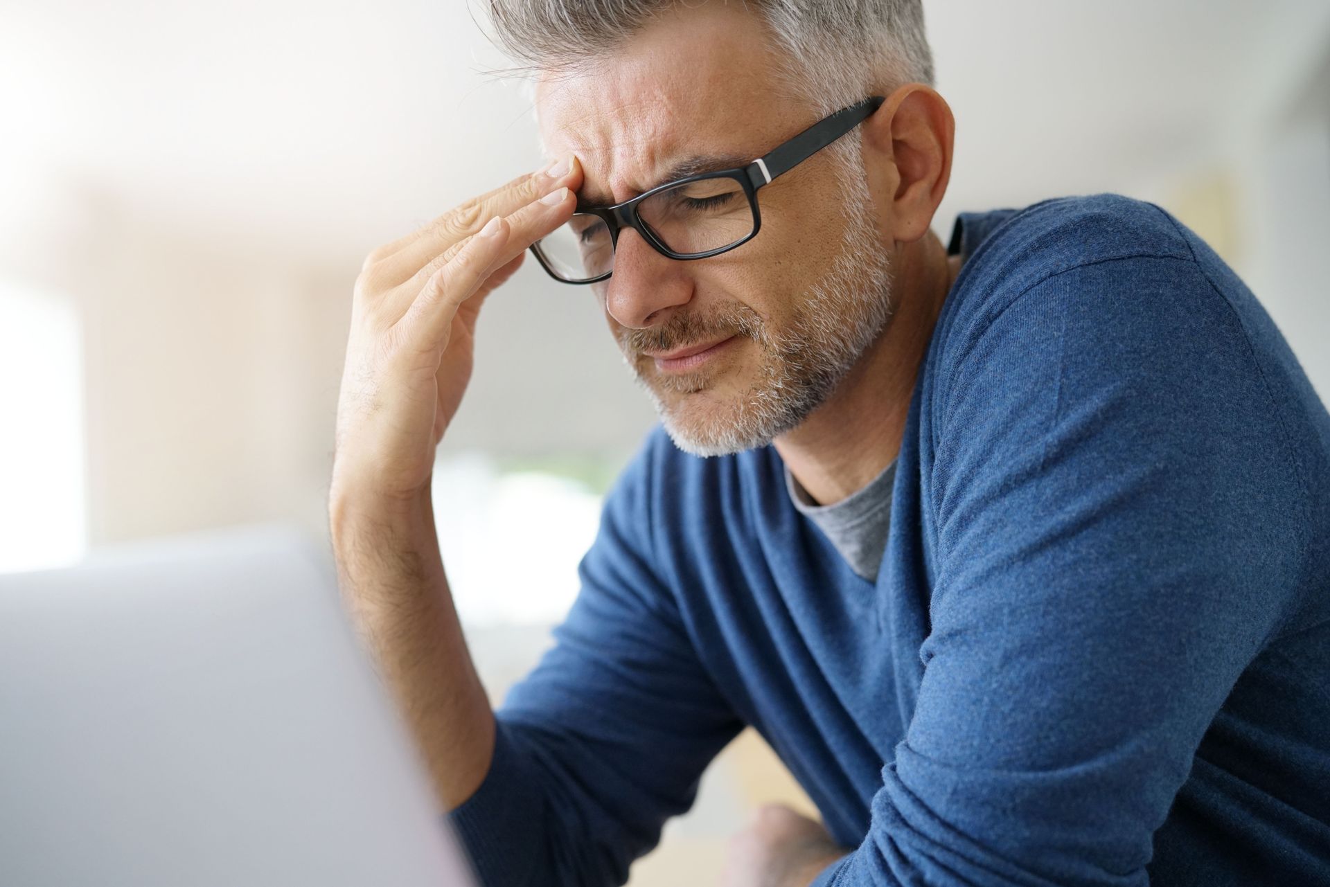 A person in a blue sweater sits at a computer, rubbing their forehead with eyes closed, appearing stressed or fatigued.