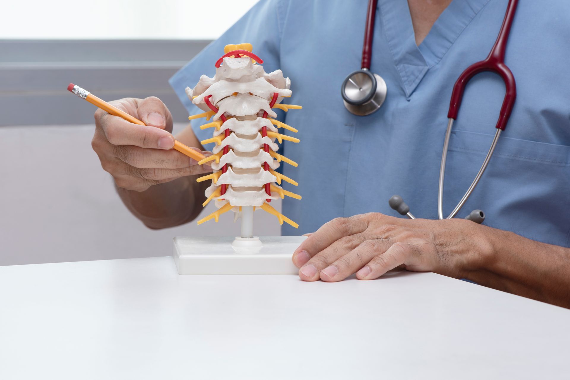 A doctor in blue scrubs with a stethoscope uses a pencil to point at a cervical spine model on desk.