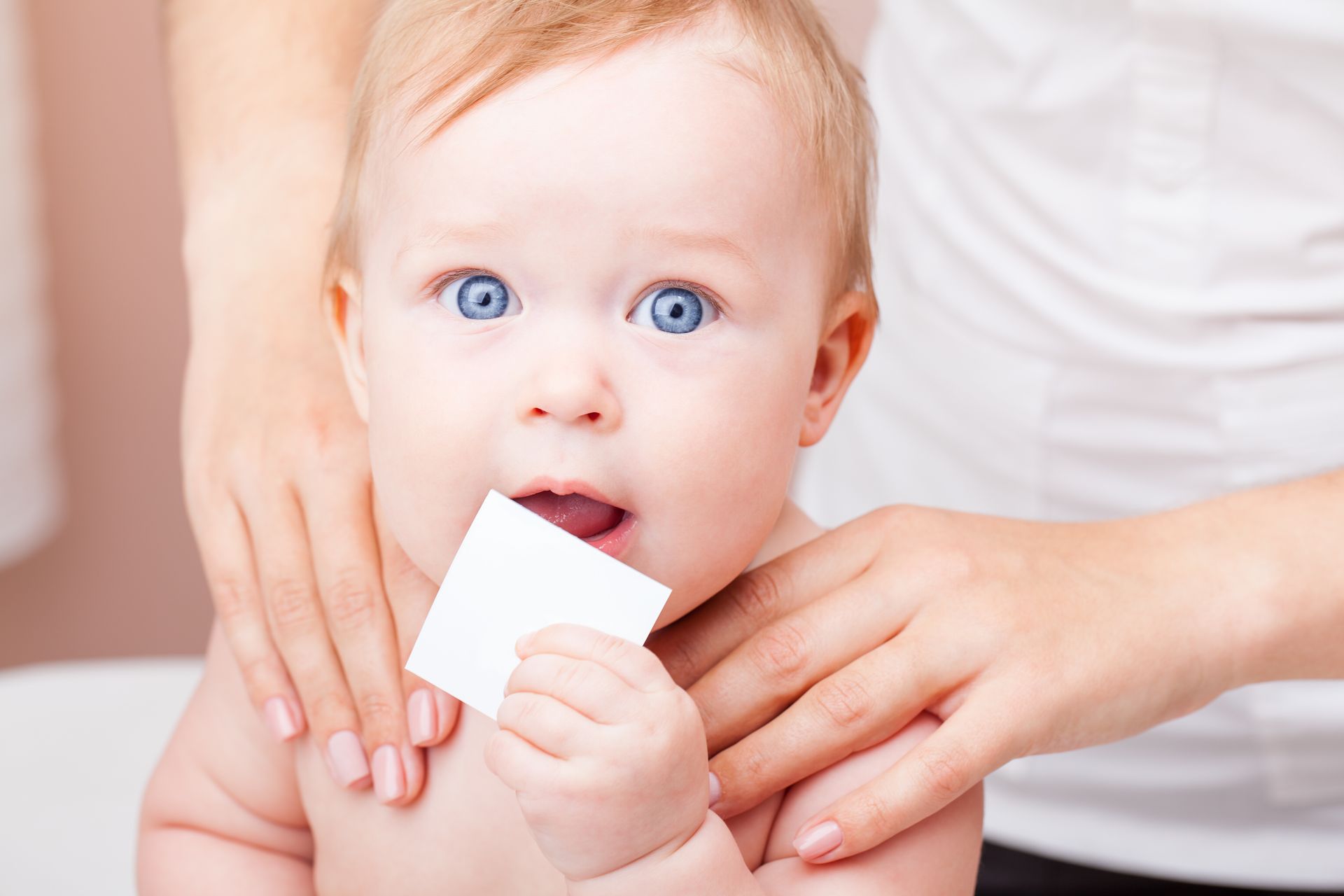 A person holds a baby, who is looking forward and holding a small white square near their mouth.