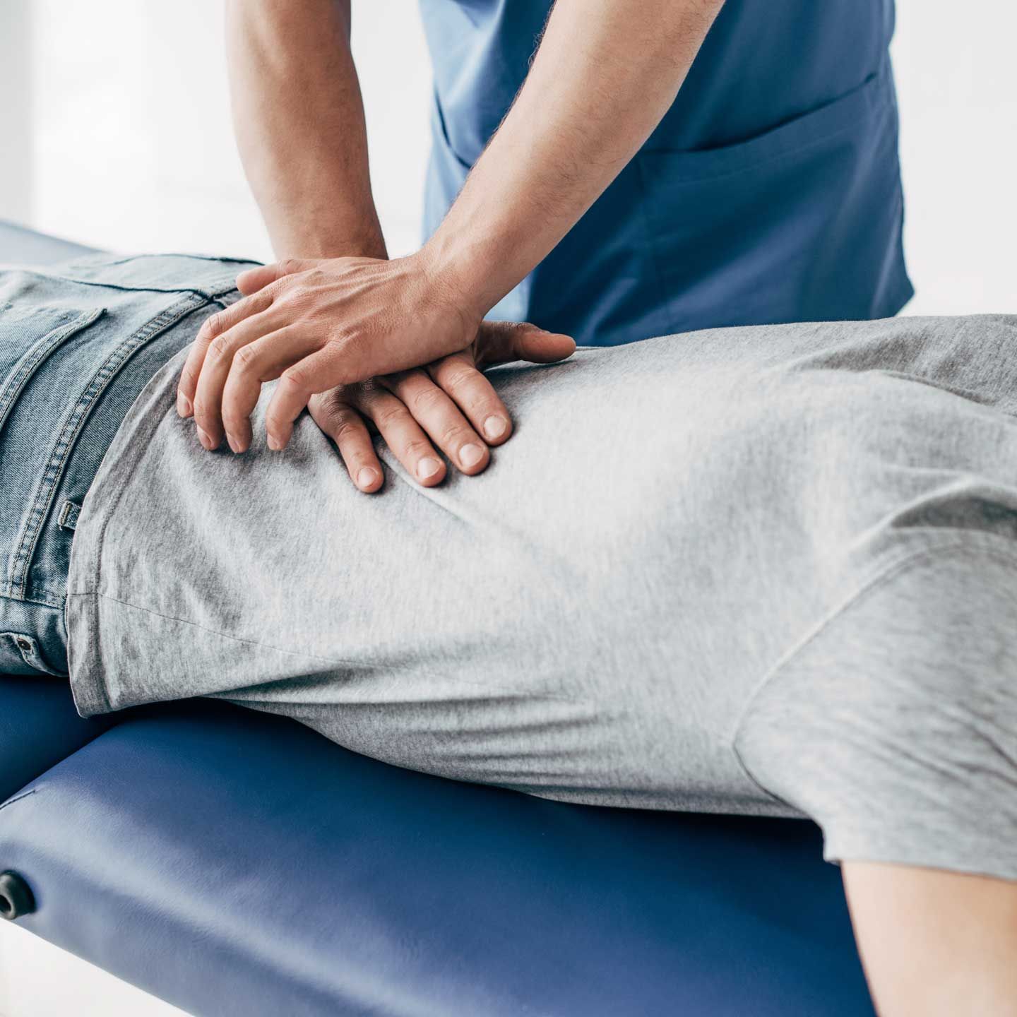 Person receiving chiropractic adjustment on a blue examination table. Chiropractor in blue scrubs with hands on patient's back. A physical therapist in blue scrubs performs a manual adjustment on the lower back of a patient lying on a clinic table.
