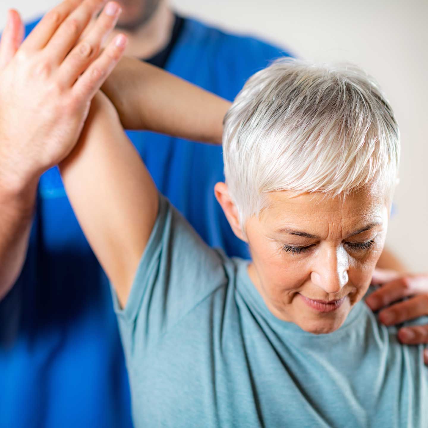 Therapist assisting a person with raising arm; indoor, white background, blue and teal clothing. A physical therapist in a blue uniform stretches the arm of a patient.