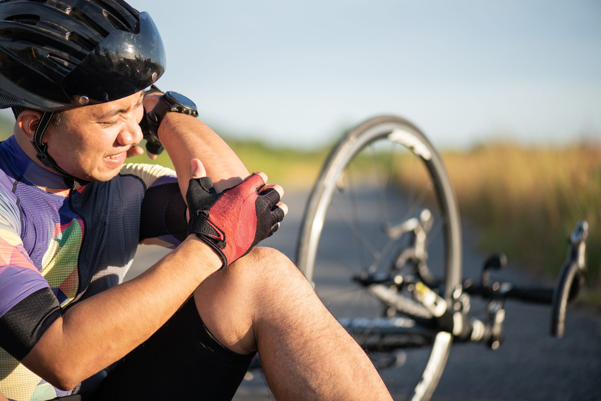 A cyclist wearing a helmet and gloves sits on a paved road, holding their arm in pain next to their bicycle.