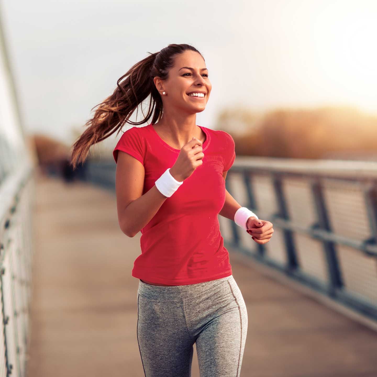 A smiling person jogs on a modern bridge during sunset, wearing a red t-shirt, grey leggings, and white wristbands.