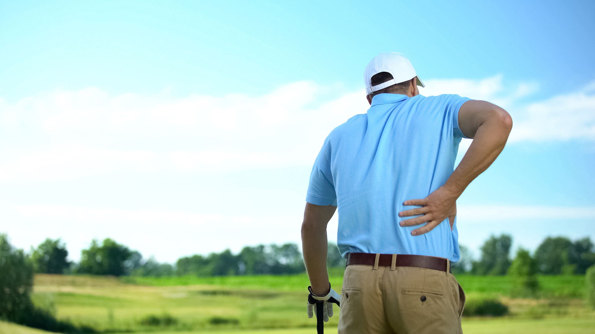 Golfer in a blue shirt and white hat standing on a course, holding their lower back due to pain.